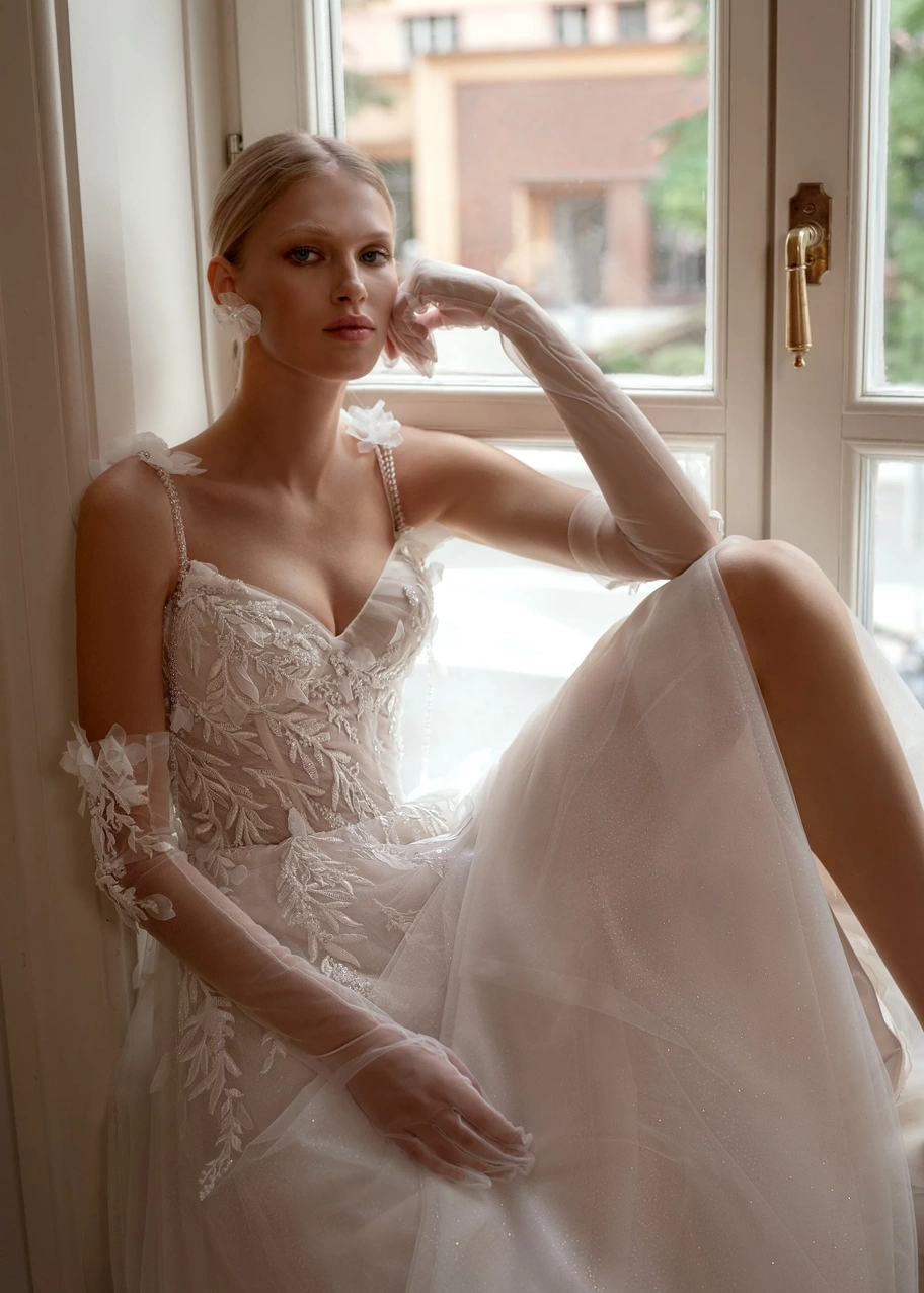 Платье «Эра» на сидящей у окна модели, с кружевным лифом и тонкими бретелями с цветами.